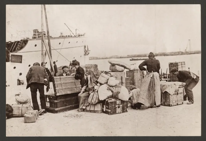 Trunks, bundles and suitcases on the quay, beside a ship’s hull. The port is not identified.
