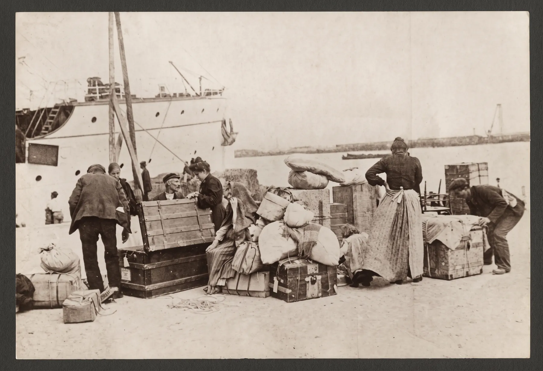 Trunks, bundles and suitcases on the quay, beside a ship’s hull. The port is not identified.