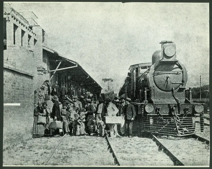 Immigrants with their luggage on a platform, beside a locomotive. The place is not identified.