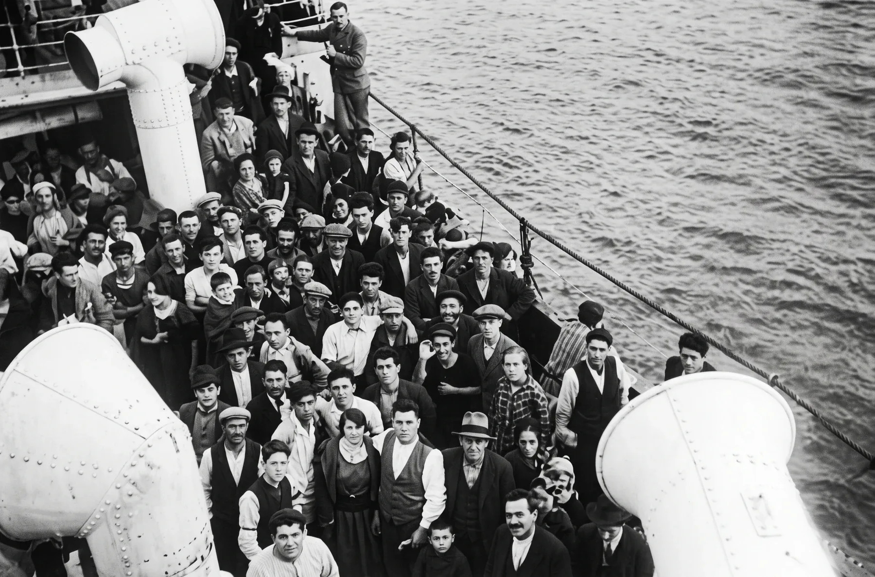 A crowd of emigrants on the deck of a liner, between two large ventilators.