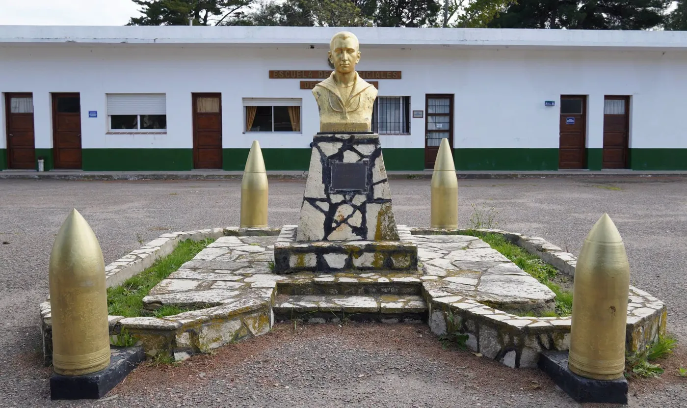 Busto dorato di Bernardi in divisa da marinaio su un piedistallo di pietra, circondato da quattro proiettili d’artiglieria dipinti d’oro, davanti a un edificio della Marina.